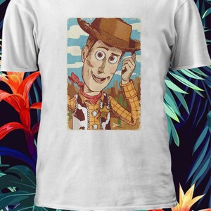 以下が含まれることがあります： トイストーリーのウッディのヴィンテージスタイルのイラストが描かれた白いTシャツ。ウッディはトレードマークのテンガロンハットとベストを着て、笑顔を見せています。イラストは茶色の枠で囲まれています。