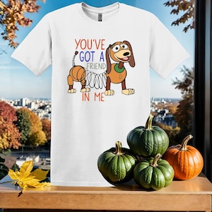 Puede incluir: Camiseta blanca con una imagen de dibujos animados de un perro con un resorte unido a su cola. El texto "You've got a friend in me" est&aacute; impreso en la camiseta.