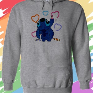 K&ouml;nnte beinhalten: Ein grauer Kapuzenpullover mit einer blauen Comicfigur, die Herzen mit bunten Buntstiften zeichnet. Die Figur hat gro&szlig;e blaue Augen und einen blauen K&ouml;rper. Der Hintergrund ist ein Regenbogen.