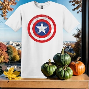 May include: White t-shirt featuring a distressed American flag design in red, white and blue. The flag is a circle with a white star in the centre.