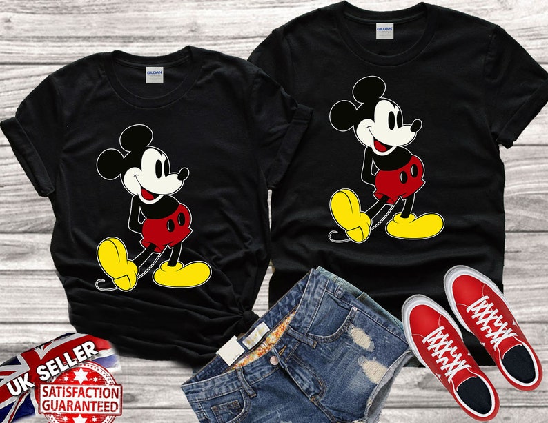 May include: Two black t-shirts with a cartoon image of Mickey Mouse in a red and yellow outfit. The shirts are laid out on a wooden surface with a pair of red sneakers and a pair of blue denim shorts.