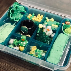 May include: A clear plastic storage box with multiple compartments filled with green and yellow themed items, including a shamrock cookie cutter, green sand, green and yellow glitter, green and yellow beads, a green rubber duck, and a green and yellow play dough.
