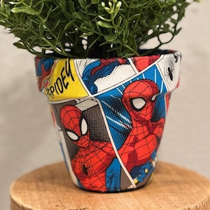 May include: A terracotta flower pot with a comic book design featuring Spiderman. The pot is decorated with red, blue, and yellow comic book panels. The pot is filled with a green plant with small white flowers.