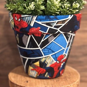 May include: A terracotta flower pot with a Spiderman design. The pot is decorated with a blue and black comic book style pattern featuring Spiderman in a red and blue suit. The pot is sitting on a wooden stool.