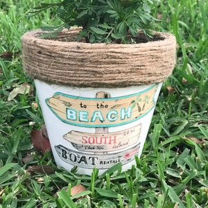 May include: A white ceramic flower pot with a brown twine rim. The pot has a painted design of a beach scene with the words "to the beach", "south", "surf club 50m", "island", and "boat rentals".