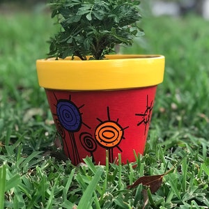 May include: A red painted flower pot with a yellow rim, decorated with black and colorful abstract designs. The pot contains a green plant with yellow flowers, set against a blurred green background.