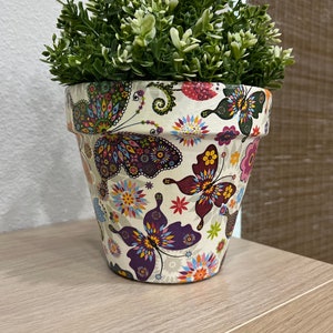 May include: A decorative flower pot with a colorful butterfly design. The pot is filled with green and white artificial plants. The pot is sitting on a light brown shelf.