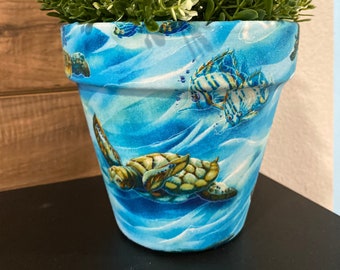 May include: A blue ceramic planter featuring a sea turtle and fish design. The planter is filled with green leafy plants.