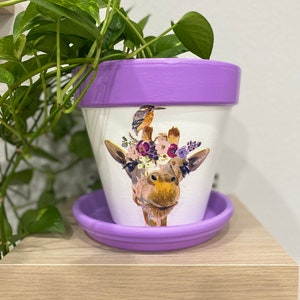 May include: Purple ceramic planter with a white giraffe design wearing a flower crown. The planter has a matching purple saucer.