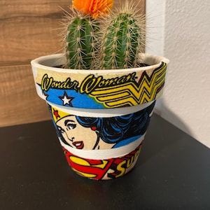 May include: A white ceramic planter with a Wonder Woman design featuring a red, yellow, and blue color scheme. The planter has a cactus with a bright orange flower growing inside.