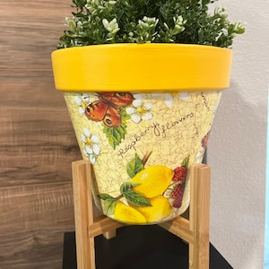 May include: A yellow ceramic planter with a floral design featuring lemons, raspberries, and butterflies. The planter is sitting on a wooden stand.