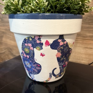 May include: A white painted flower pot with a blue rim and a floral cat design. The pot contains a green plant. The cat design features blue and pink floral patterns and two pink butterflies.