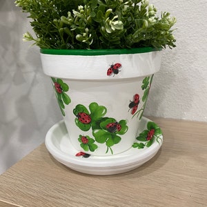 May include: White ceramic flower pot with a green rim, decorated with green four-leaf clovers and red ladybugs. The pot has a matching white saucer.