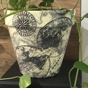 May include: A cream-colored ceramic planter with a vintage world map design. The planter features a compass rose and multiple maps of the world. A green plant is growing in the planter.