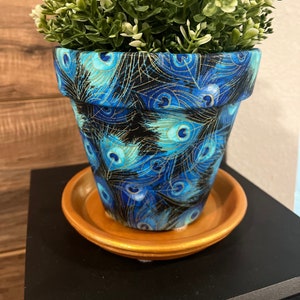 May include: A blue and gold peacock feather patterned terracotta planter with a green artificial plant inside and a brown saucer underneath.