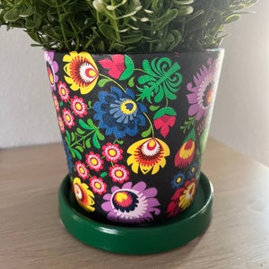 May include: A black planter with a green saucer. The planter is decorated with colorful folk art flowers. A small green plant is in the planter.