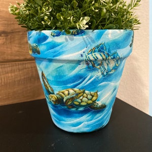 May include: A blue ceramic planter with a sea turtle and fish design. The planter is filled with green leafy plants.