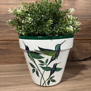 May include: A white ceramic flower pot with a green rim and a design of three hummingbirds and leaves. The pot is filled with green and white faux greenery. The pot is sitting on a wooden surface.