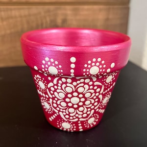 May include: A small terracotta pot painted in a vibrant pink with a white mandala design. The pot is perfect for small plants or succulents.