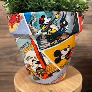 May include: A decorative flower pot featuring a collage of vintage Disney cartoon images, including Mickey Mouse. The pot is filled with green foliage and sits on a small wooden stool. The pot's design includes various colors.