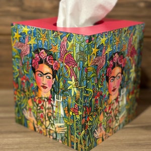May include: A pink tissue box cover with a colorful illustration of Frida Kahlo surrounded by flowers and birds. The background is a dark blue with gold stars.
