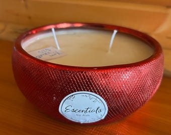 May include: A red, textured bowl-shaped candle with two wicks. The candle has a light-colored wax and a label that reads "Escentials by Ash". The candle sits on a wooden surface.