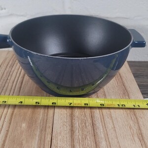 May include: A blue fondue pot with a handle and two side handles. The pot is sitting on a wooden surface. A yellow tape measure is placed next to the pot, showing a measurement of approximately 11 inches.
