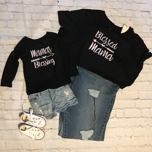 May include: Two black long-sleeve shirts with white text, one reading Blessed Mama and the other Mamas Blessing, each featuring an arrow graphic. Included are a pair of light blue distressed denim shorts, blue distressed jeans, and white sneakers.