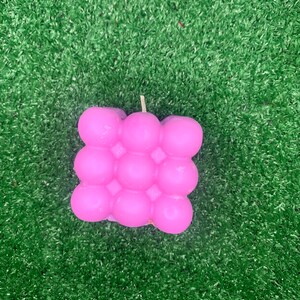 Könnte beinhalten: Eine rosa Blasenkerze in quadratischer Form. Die Kerze hat eine strukturierte Oberfläche mit mehreren runden Noppen.