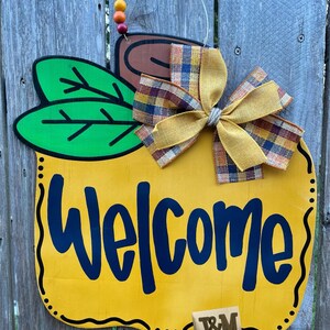 May include: A yellow pumpkin-shaped welcome sign with a brown stem, green leaves, and a plaid and yellow bow. The sign says "Welcome" in black lettering and has a brown "J&M" monogram on the bottom.