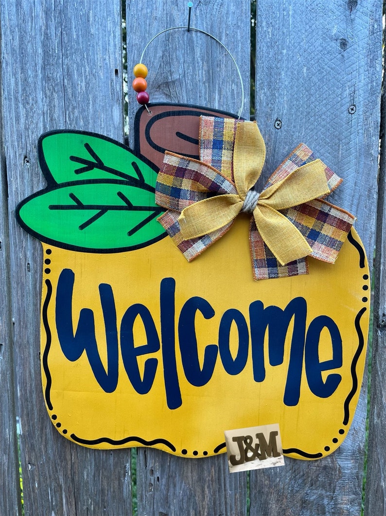 May include: A yellow pumpkin-shaped welcome sign with a brown stem, green leaves, and a plaid and yellow bow. The sign says "Welcome" in black lettering and has a brown "J&M" monogram on the bottom.