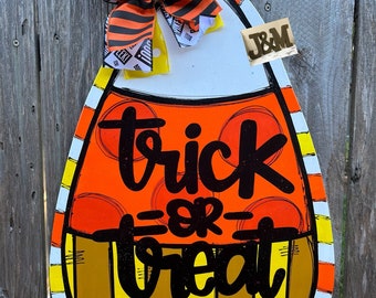 May include: A wooden candy corn door hanger with a black and orange striped bow. The door hanger is painted with orange, yellow, and white stripes and has the words "trick or treat" written in black.