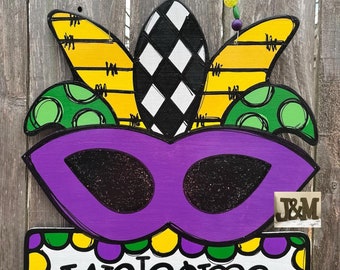 May include: Purple and green Mardi Gras mask welcome sign with yellow and black fleur-de-lis accents. The sign says "Welcome" in white lettering.