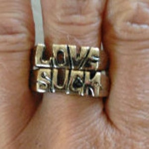 May include: Two gold-tone rings with the words "LOVE" and "SUCK" engraved on them. The rings are stacked on top of each other.