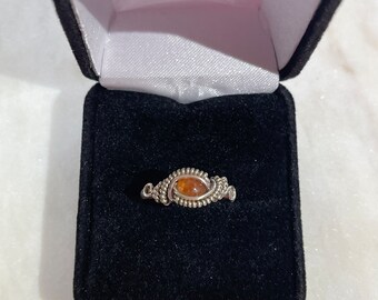 May include: A silver ring with a brown gemstone set in a wire-wrapped design. The ring is displayed in a black velvet ring box.
