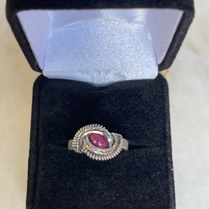 May include: A silver ring with a deep red gemstone set in a wire wrapped design. The ring is displayed in a black velvet ring box.