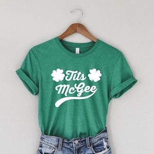 May include: A green t-shirt with rolled-up sleeves and the text "Tits McGee" in white, along with two four-leaf clovers. The shirt is paired with denim shorts, hanging on a wooden hanger against a neutral background.