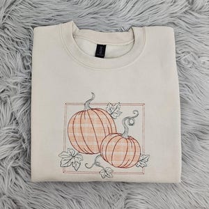 May include: Cream-colored sweatshirt featuring an embroidered design of two pumpkins with a plaid pattern, framed by a rectangular border and leaves. The pumpkins are in shades of orange and brown, with black outlines. The sweatshirt is folded on a gray, fuzzy surface.
