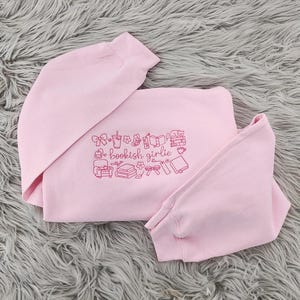 May include: A light pink long-sleeve sweatshirt featuring dark pink embroidered line art of books, a chair, a cat, a bow, a drink, and a bookmark. The text "bookish girlie" is centered within the design, which is surrounded by small floral accents.