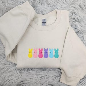 May include: A light beige sweatshirt with a row of embroidered pastel-colored bunny shapes. The bunnies are yellow, pink, light pink, purple, blue, and green.