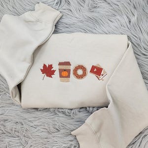 May include: A beige sweatshirt with an embroidered design of a fall-themed scene. The design includes a maple leaf, a cup of coffee with a pumpkin inside, a donut, and a book with a heart on the cover.