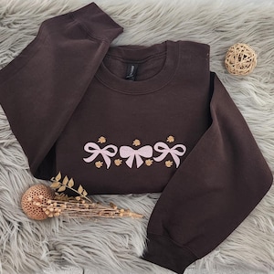 May include: A brown sweatshirt with a row of pink bows embroidered on the front. The bows are outlined in gold thread.