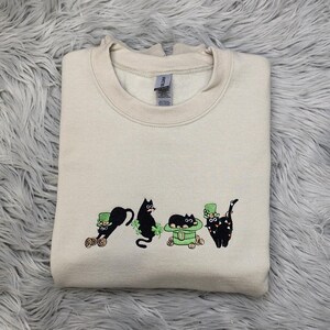 May include: A cream-colored sweatshirt with an embroidered design of five black cats wearing green hats and holding shamrocks. The cats are arranged in a line, with the first cat holding a pot of gold.