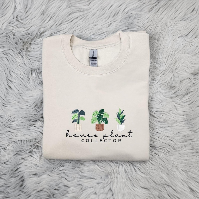 Puede incluir: Una sudadera de color crema con un dise&ntilde;o bordado de tres plantas en maceta y el texto "house plant collector".