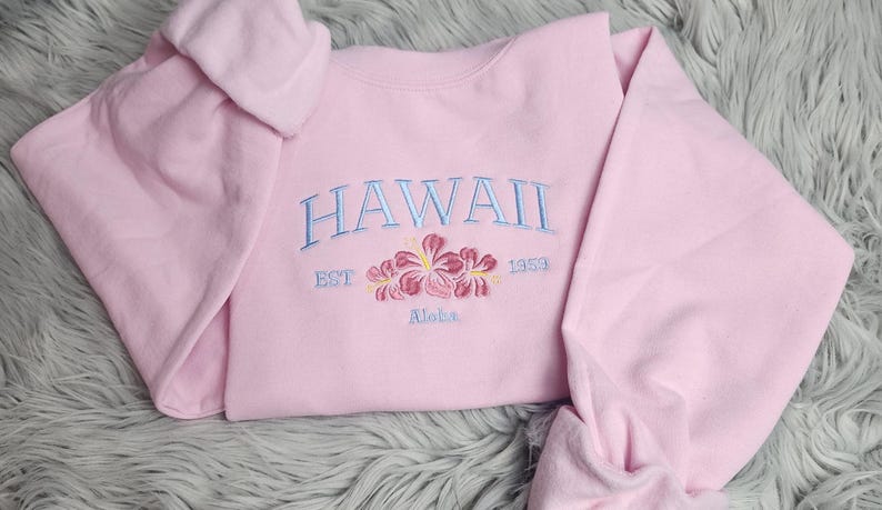 May include: Pink sweatshirt with the text "HAWAII EST 1988 Aloha" embroidered in blue and pink. The sweatshirt features a floral design with pink hibiscus flowers.