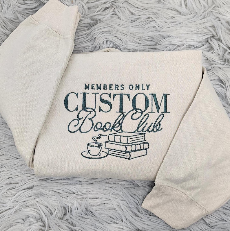 May include: A cream-colored sweatshirt with the text "MEMBERS ONLY CUSTOM BOOK CLUB" in dark blue lettering. A stack of books and a cup of coffee are illustrated below the text.