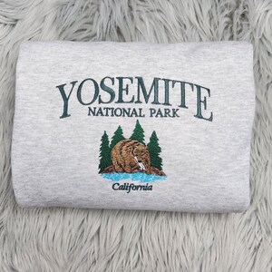 May include: A gray sweatshirt with an embroidered design featuring the words "Yosemite National Park" and a bear in a forest setting. The text "California" is also embroidered below the bear.