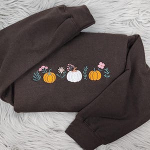 May include: A dark brown sweatshirt with embroidered pumpkins and floral accents. The design features two orange pumpkins, a white pumpkin, and various pink and green floral elements. The sweatshirt is folded on a light gray, fuzzy surface.