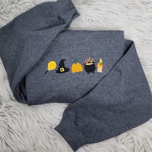 May include: Dark gray sweatshirt with embroidered Halloween-themed designs. The design includes a lollipop, witch hat, pumpkin, cauldron, and candle. The sweatshirt is folded on a white, fluffy surface.