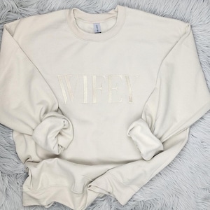 May include: A cream-colored sweatshirt with the word "WIFEY" embroidered in a light beige color.